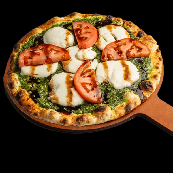 HOF Flatbreads pesto flatbread with fresh mozzarella, sliced tomatoes, and balsamic glaze on a wooden peel.